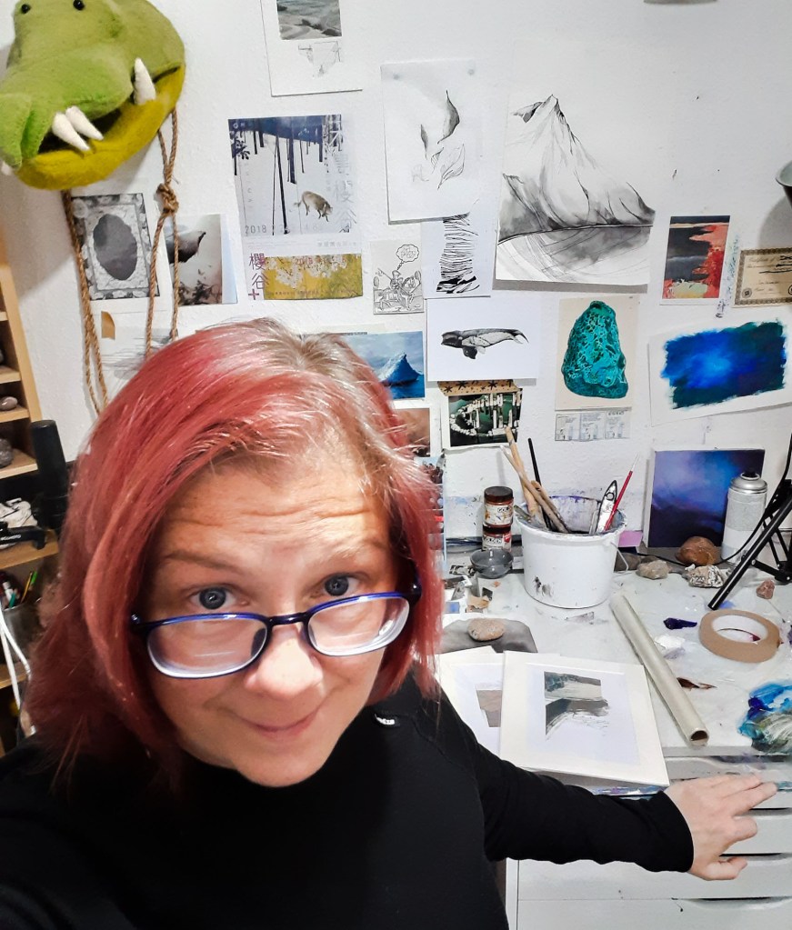 Photo of the artist looking at the camera, studio table and wall of art in the background